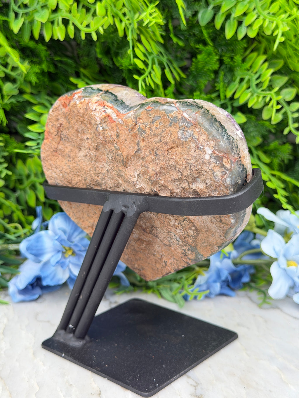 A heart-shaped rock is mounted on a black metal bracket, surrounded by lush green foliage and blue flowers.
