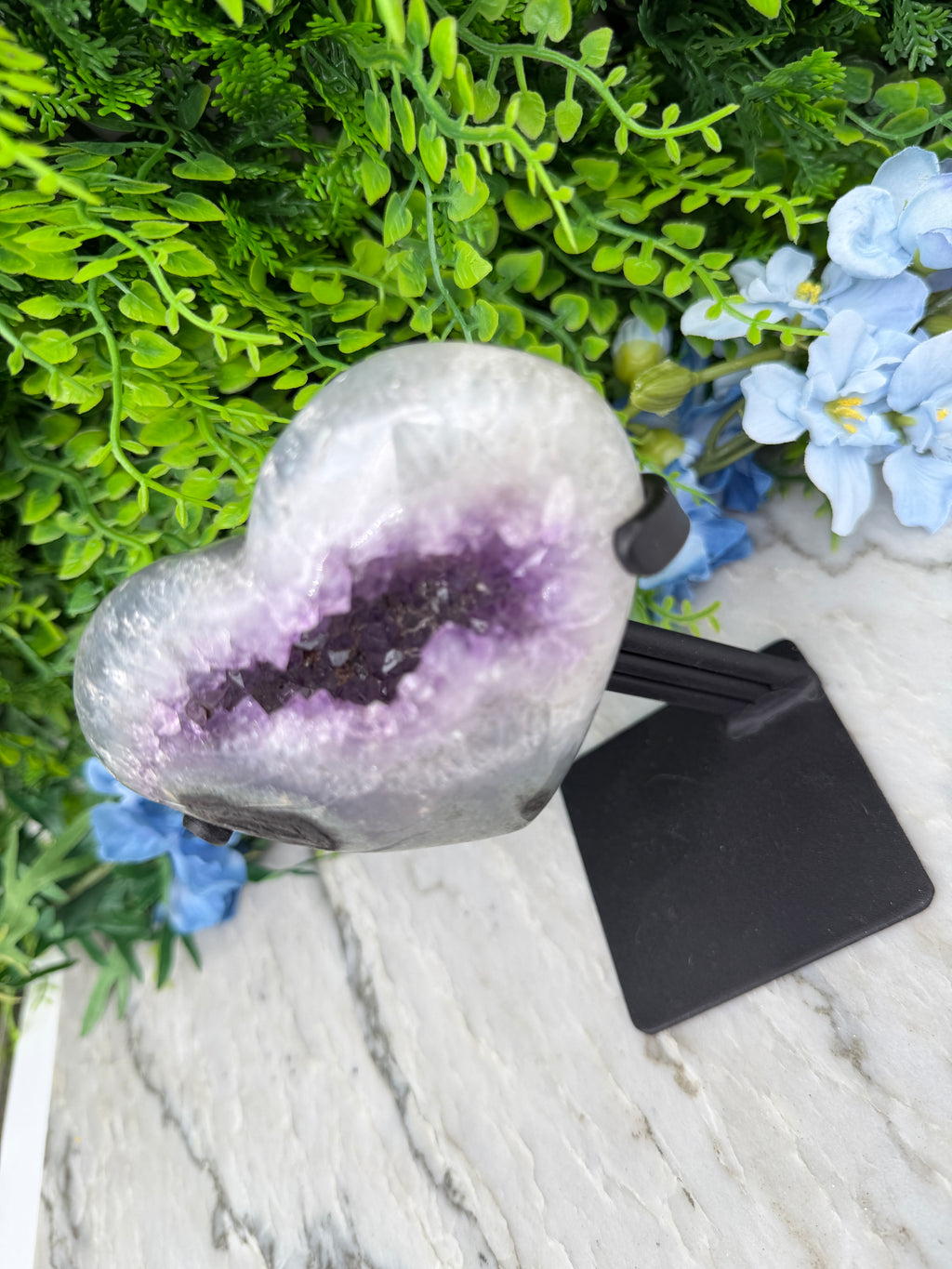 a heart-shaped amethyst geode with purple crystals, surrounded by green foliage and blue flowers.