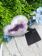 a heart-shaped amethyst geode with purple crystals, surrounded by green foliage and blue flowers.