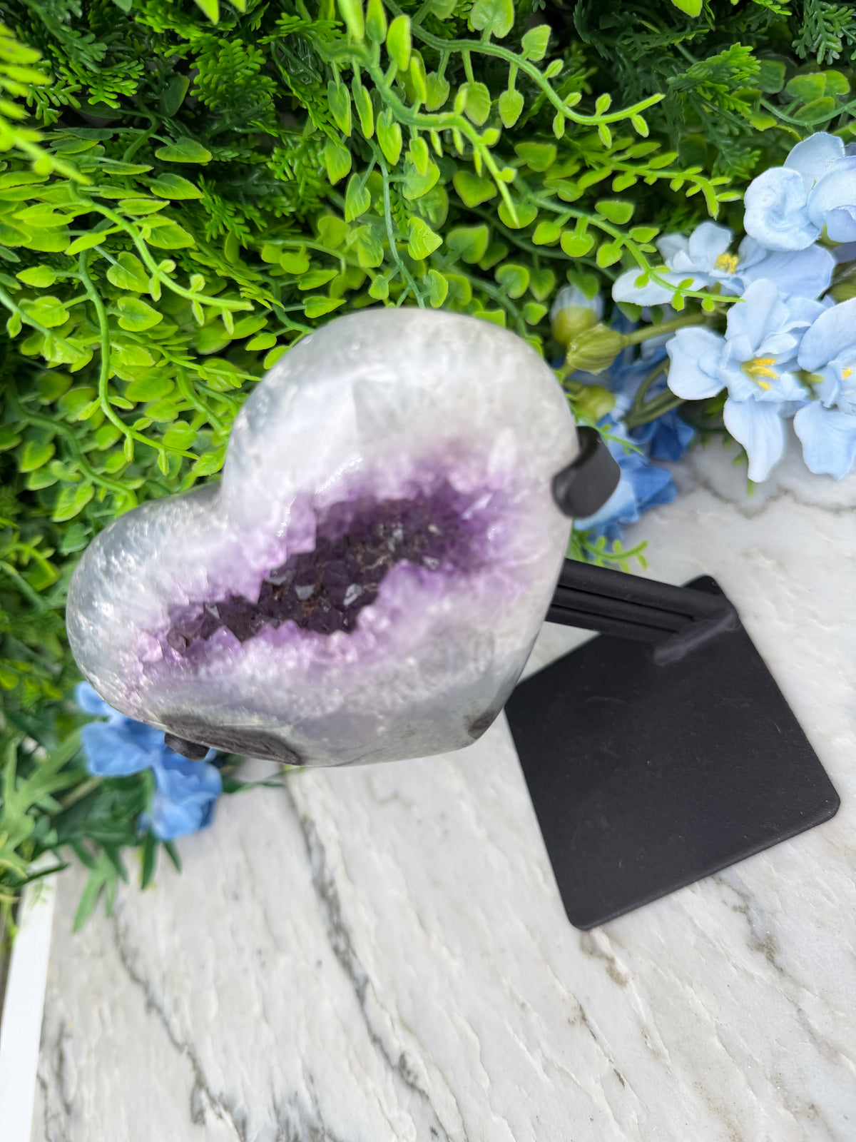a heart-shaped amethyst geode with purple crystals, surrounded by green foliage and blue flowers.