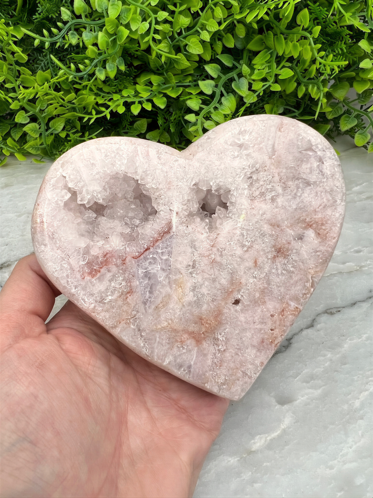A hand is holding a heart-shaped geode with a pinkish-white color, set against a background of green foliage.