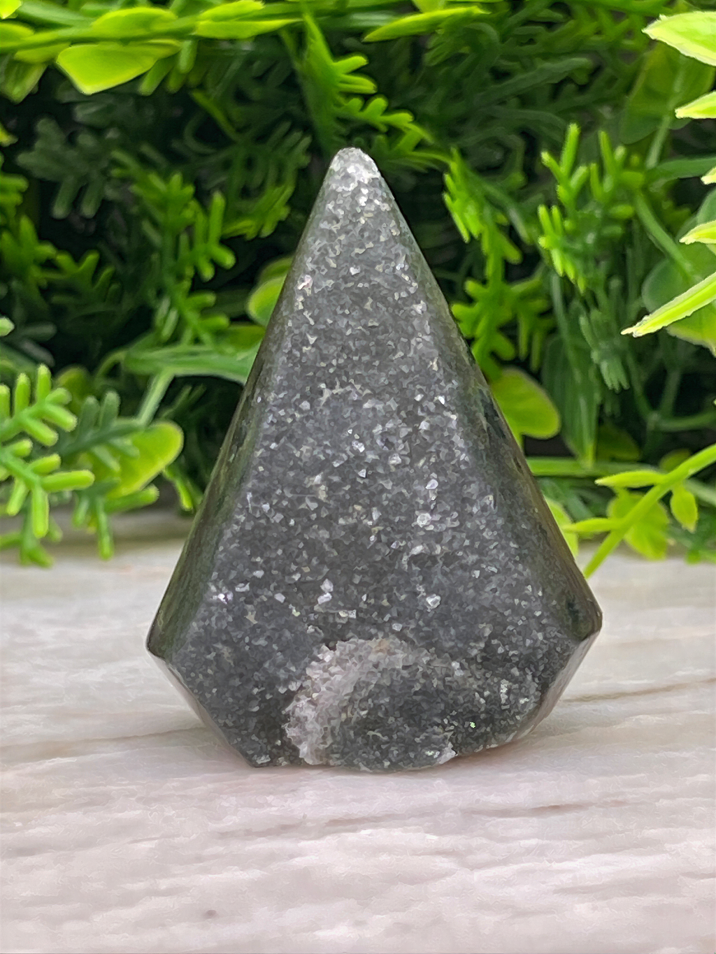 A triangular-shaped, black-colored stone or gemstone is placed on a white surface, surrounded by green foliage.