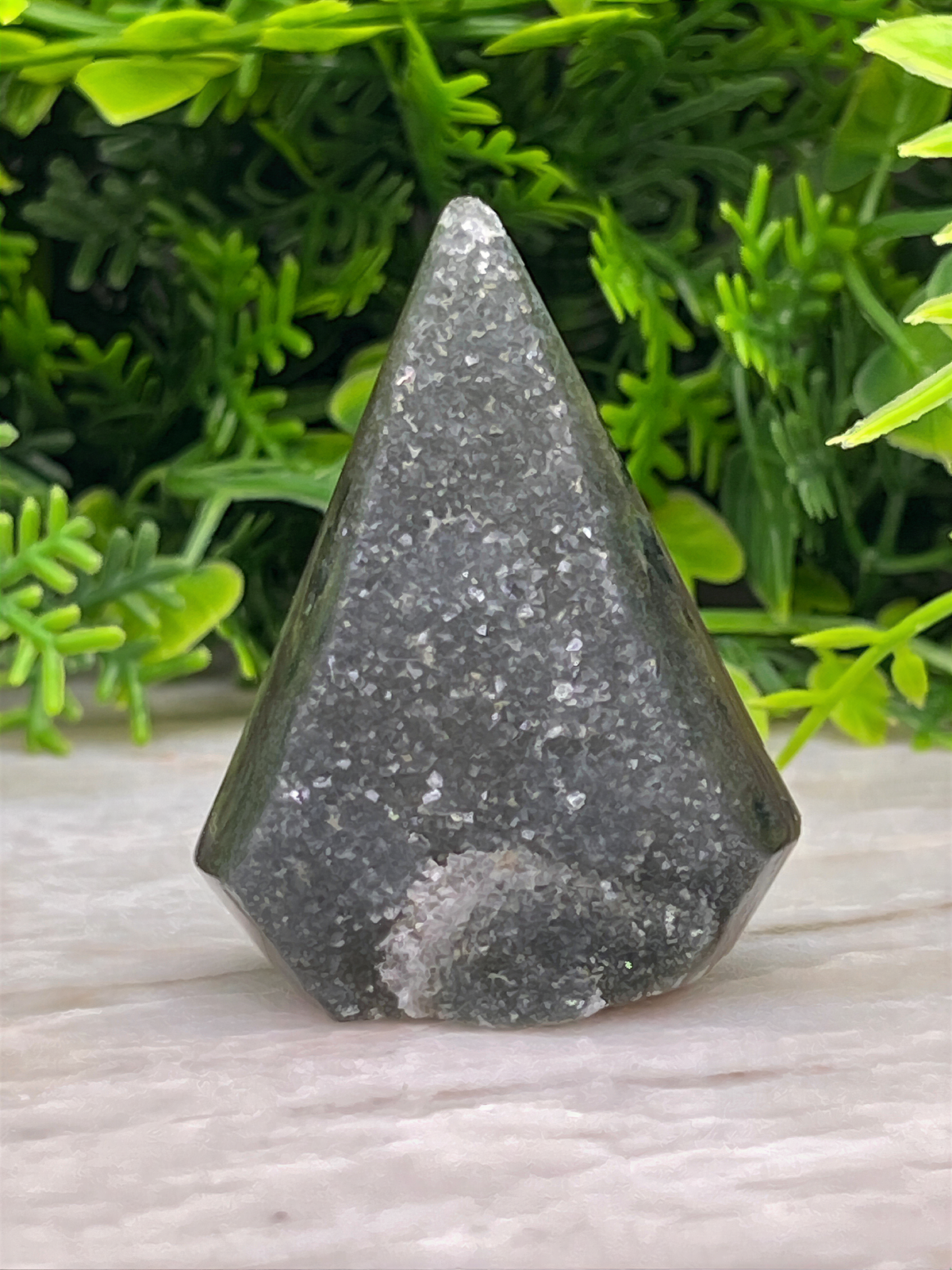 A triangular-shaped, black-colored stone or gemstone is placed on a white surface, surrounded by green foliage.