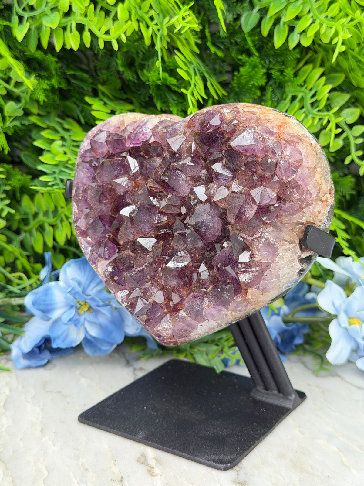 A heart-shaped crystal formation with purple crystals is displayed against a backdrop of green foliage and blue flowers.