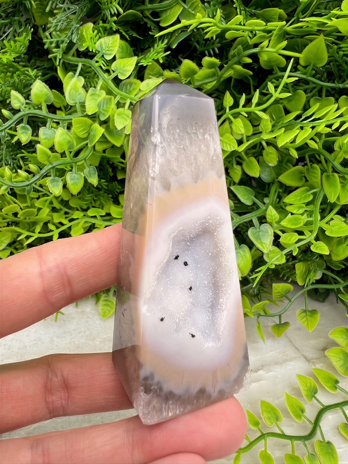A hand is holding a clear, triangular-shaped crystal with a white and gray color pattern, set against a background of green foliage.