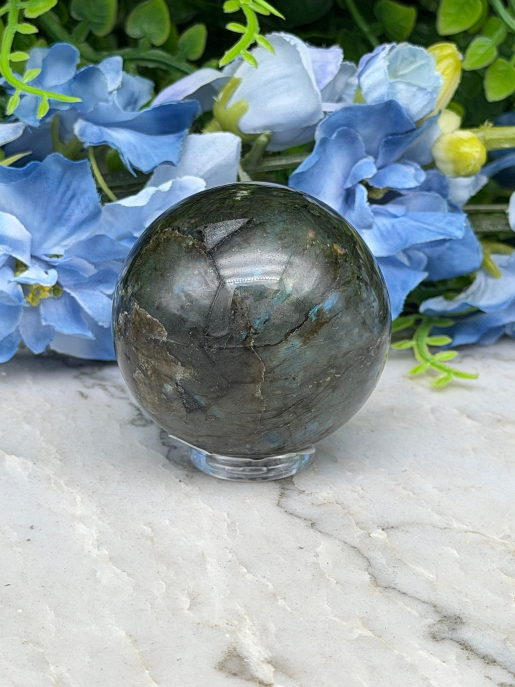 A close-up image of a dark green crystal ball surrounded by blue flowers, with a white marble surface in the background.