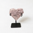A heart-shaped object made of a textured material—possibly stone or metal—displayed on a black stand against a plain white background.