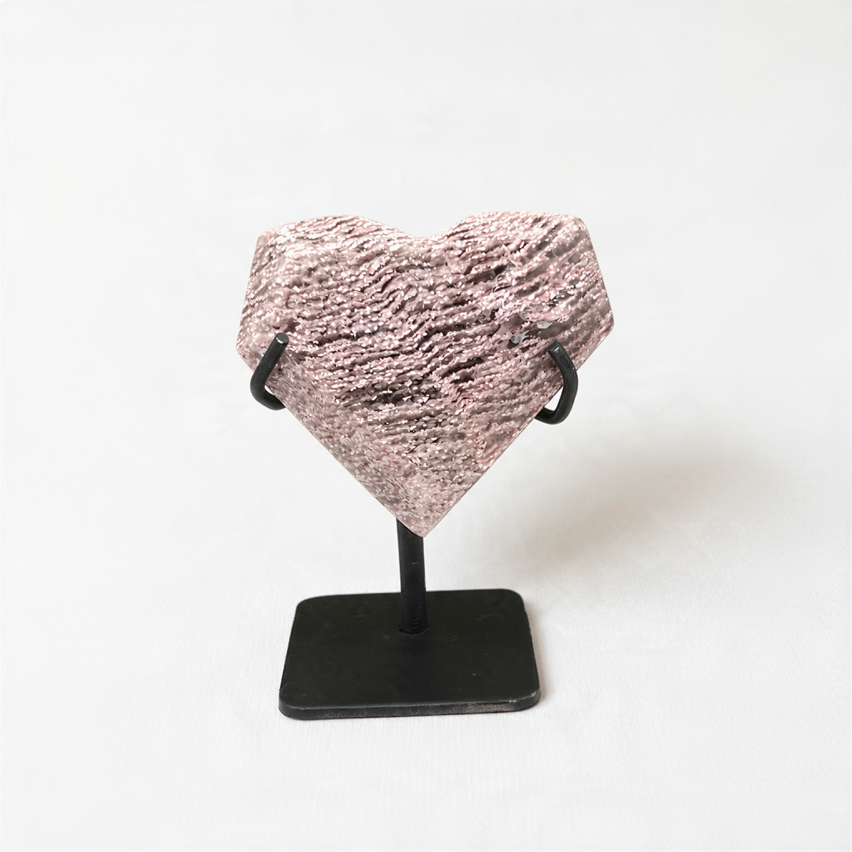 A heart-shaped object made of a textured material—possibly stone or metal—displayed on a black stand against a plain white background.
