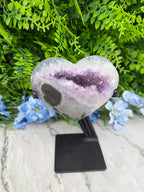 A heart-shaped amethyst geode is displayed on a black stand, surrounded by lush green foliage and blue flowers.