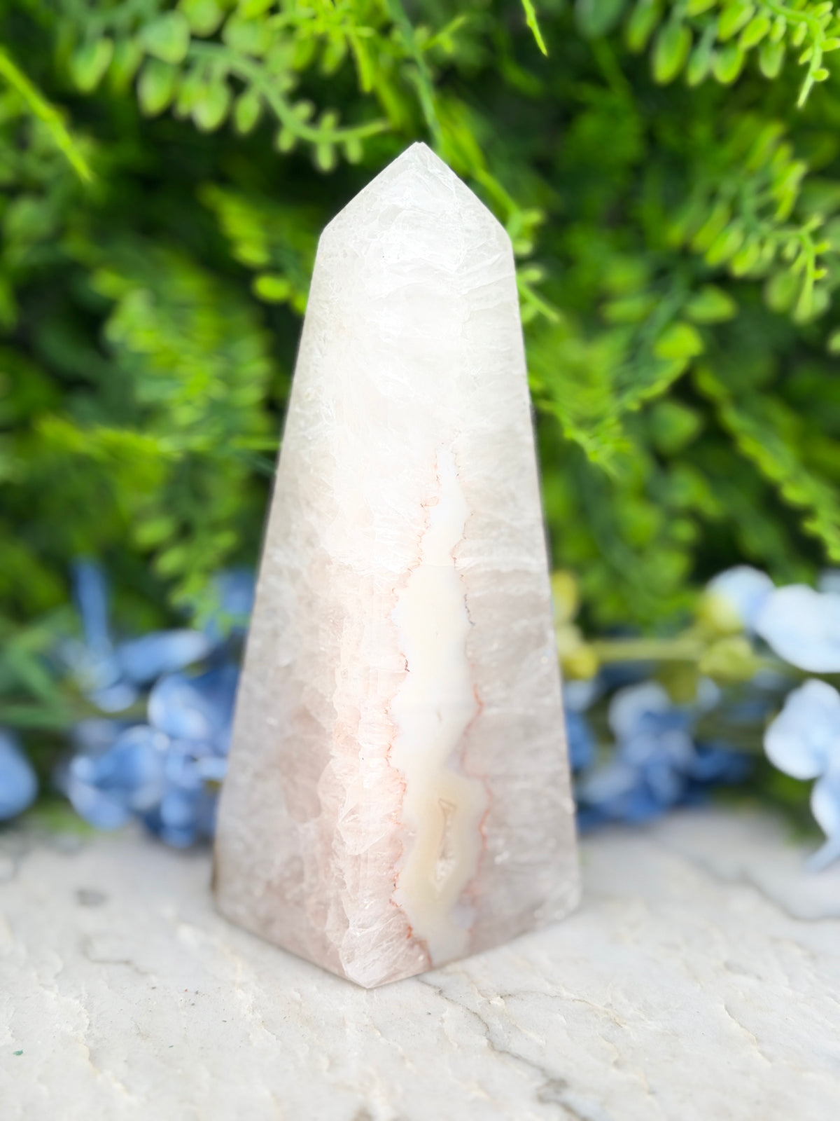 A white, triangular crystal sits on a white surface, surrounded by lush green foliage and blue flowers.
