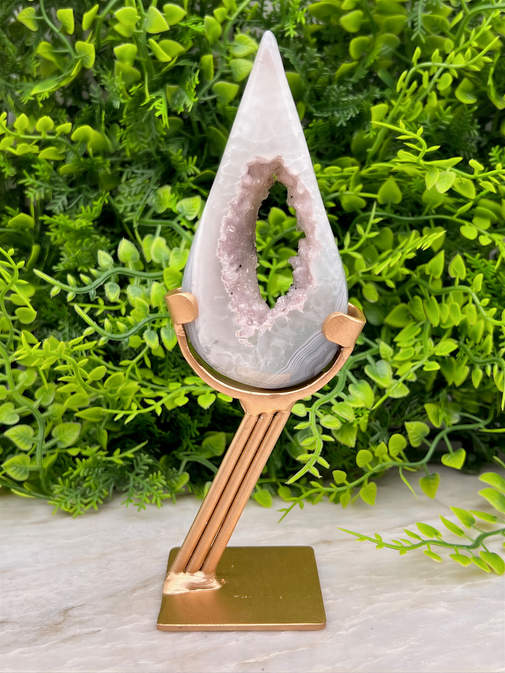 A white geode with a pinkish-purple interior is displayed on a gold stand, surrounded by lush green foliage.