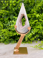 A white geode with a pinkish-purple interior is displayed on a gold stand, surrounded by lush green foliage.