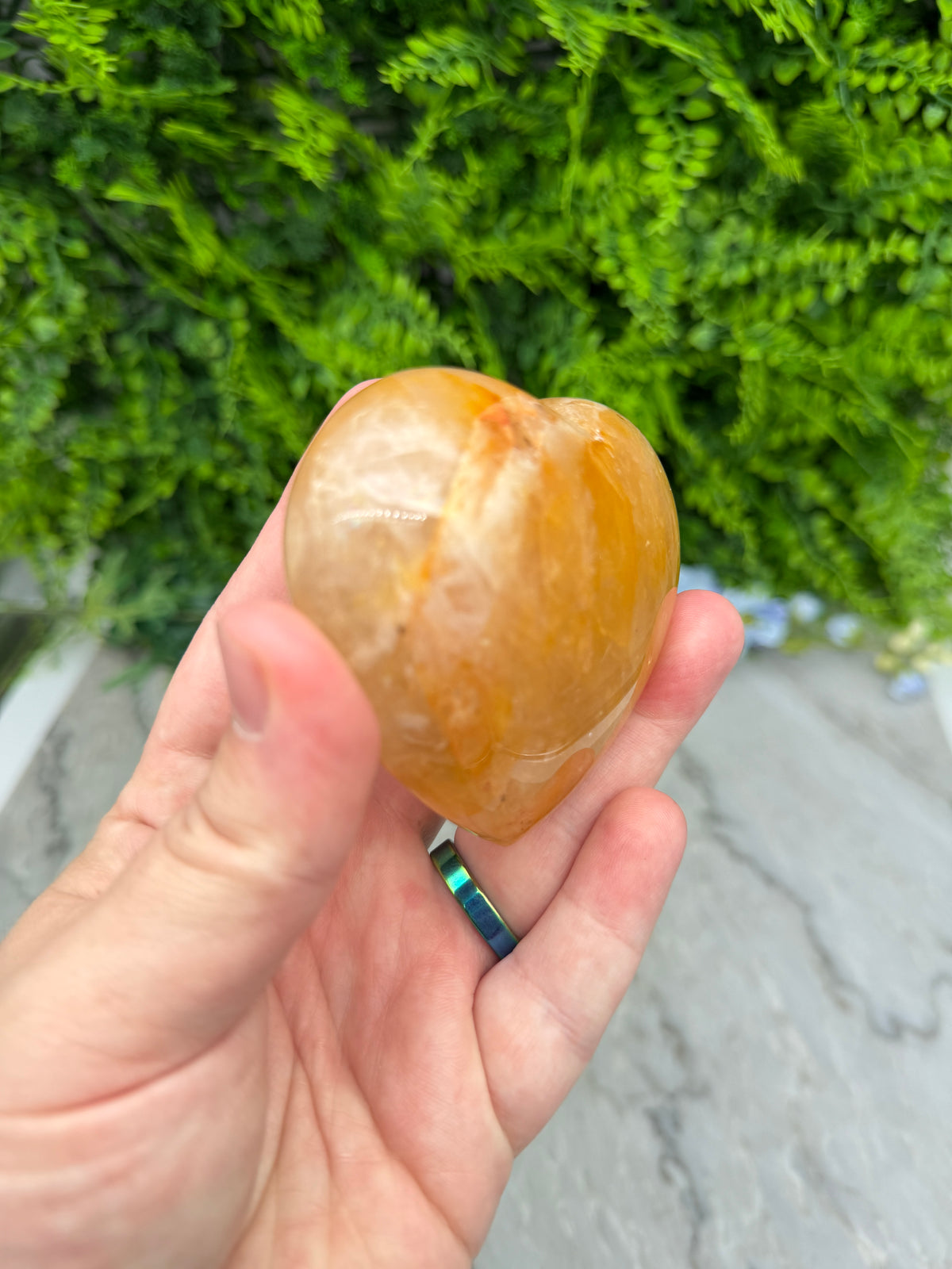 A hand holding a small, round, orange-colored stone or gemstone against a backdrop of lush green foliage.