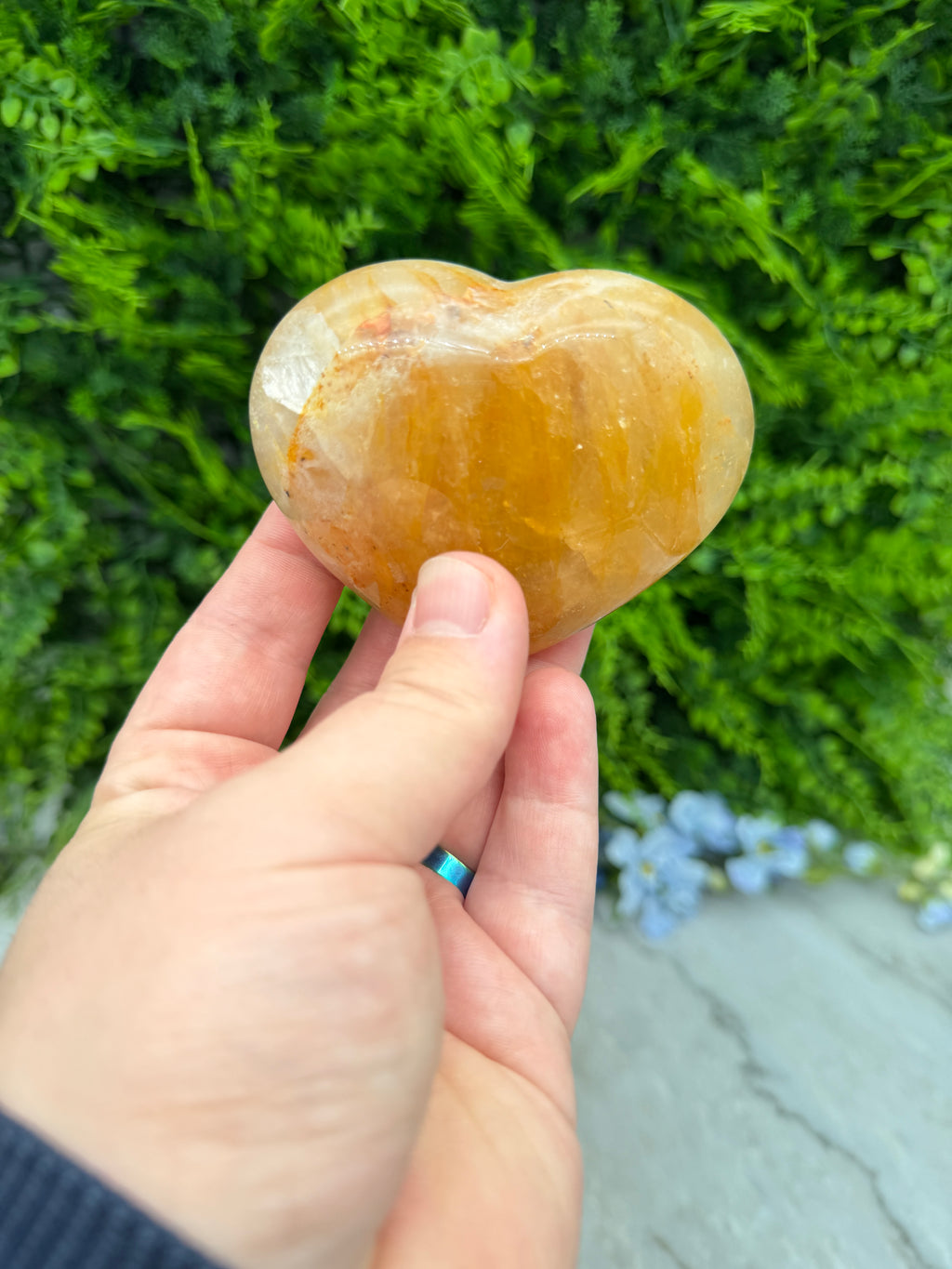 A hand holding a heart-shaped crystal against a backdrop of green foliage.