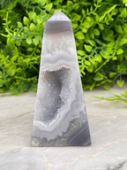 A gray and white geode-like rock formation with a pointed top sits on a white surface, surrounded by lush green foliage.