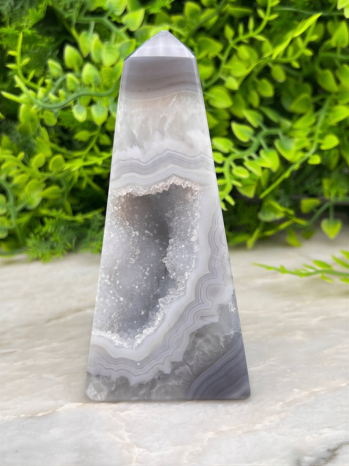 A gray and white geode-like rock formation with a pointed top sits on a white surface, surrounded by lush green foliage.