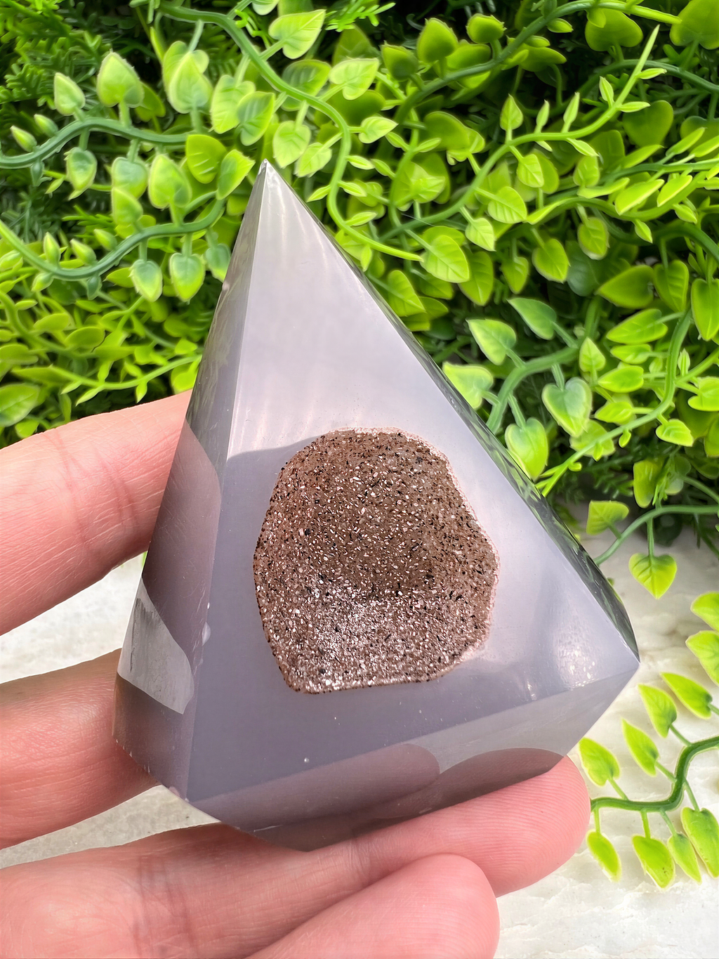 A hand holding a triangular, gray-colored object with a small, brown-colored stone or mineral embedded in it, set against a background of green foliage.