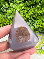A hand holding a triangular, gray-colored object with a small, brown-colored stone or mineral embedded in it, set against a background of green foliage.