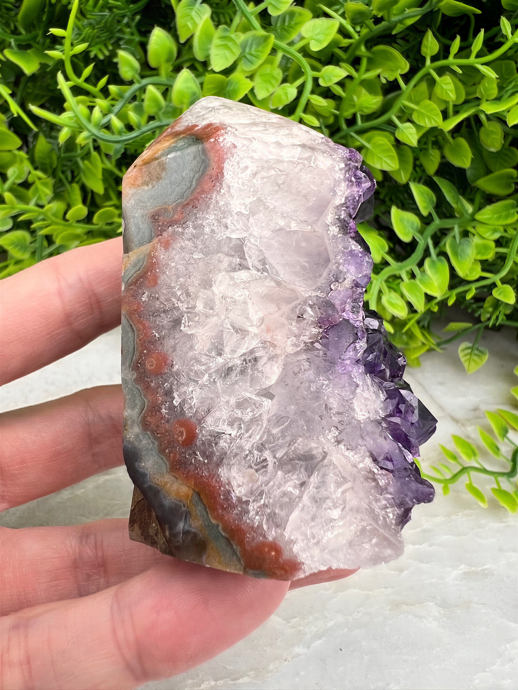 A hand is holding a piece of amethyst, a purple crystal, against a backdrop of green foliage.