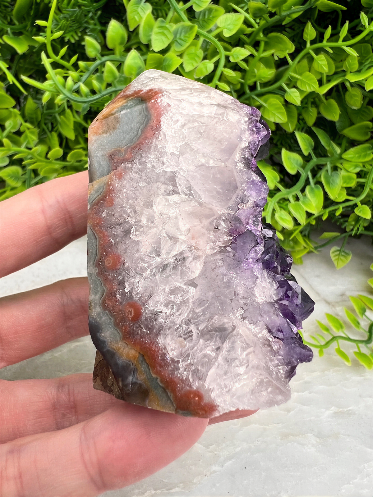 A hand is holding a piece of amethyst, a purple crystal, against a backdrop of green foliage.