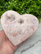 A hand is holding a heart-shaped geode with crystals inside, set against a backdrop of green foliage.