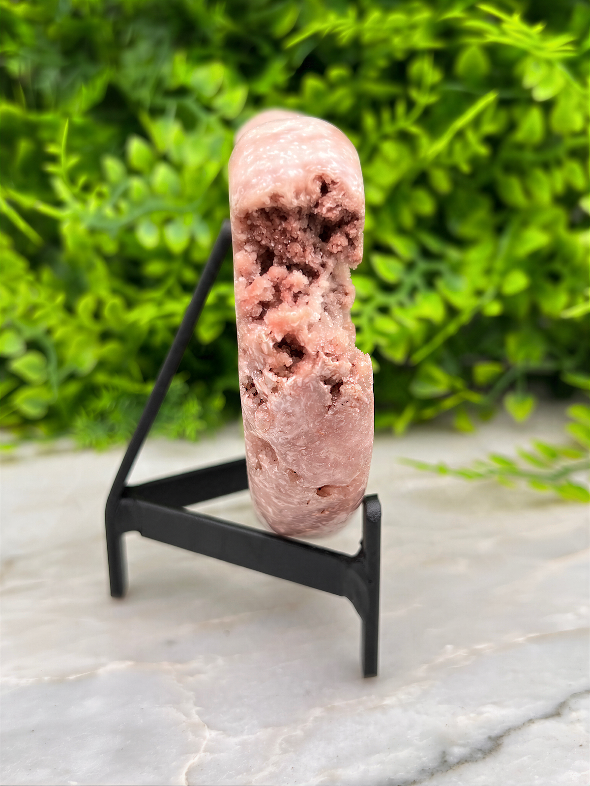 A pink crystal or rock formation is displayed on a black stand, against a blurred green background.
