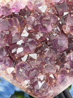 a close-up view of a crystal formation, with numerous small, clear crystals arranged in a pattern. The crystals appear to be purple in color, and they are set against a blurred background.