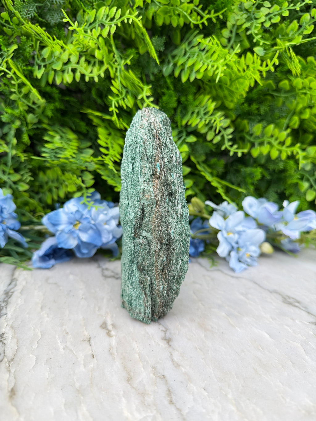 A close-up view of a green rock with a rough texture, set against a backdrop of lush green foliage and delicate blue flowers.