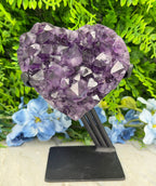 A heart-shaped piece of purple amethyst crystal is displayed against a backdrop of green foliage and blue flowers.