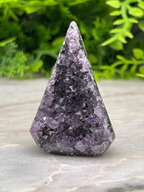 A triangular piece of amethyst crystal with a dark purple hue sits on a white surface, surrounded by lush green foliage.