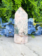 A jade crystal with a greenish hue sits on a marble surface, surrounded by blue flowers.
