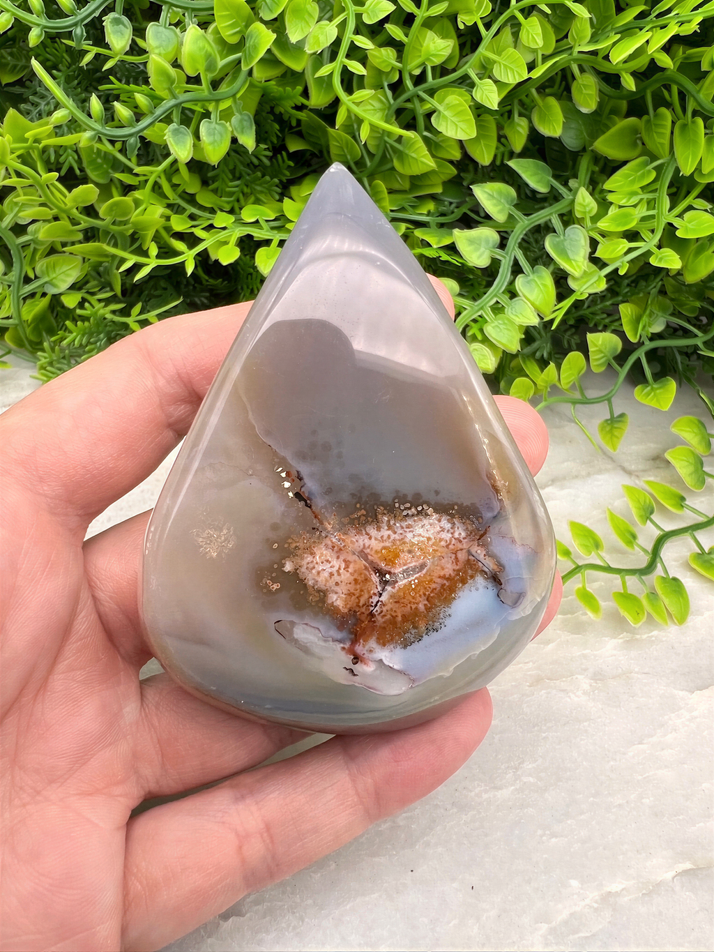 A hand is holding a teardrop-shaped agate stone with a brownish speckled surface, set against a backdrop of green foliage.