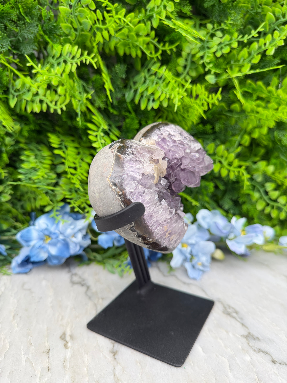 a large purple crystal surrounded by green foliage and blue flowers, mounted on a black stand.