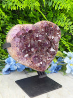 A large, heart-shaped crystal formation with numerous small purple crystals embedded in it, set against a backdrop of green foliage and blue flowers.