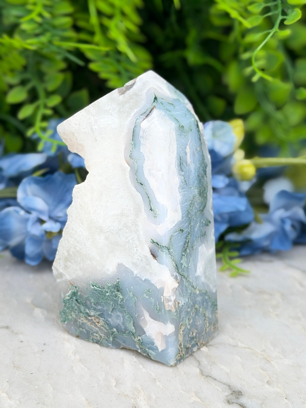 A white and green jadeite crystal sits on a white surface, surrounded by blue flowers.