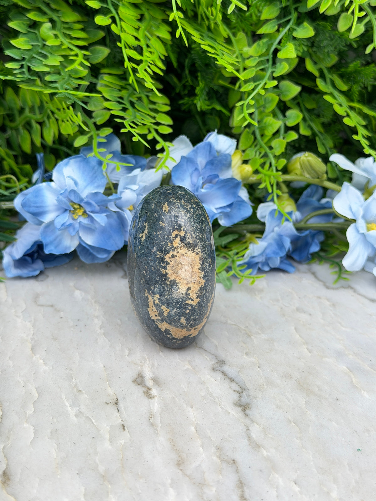A black rock with a speckled surface rests on a white surface, surrounded by vibrant blue flowers.