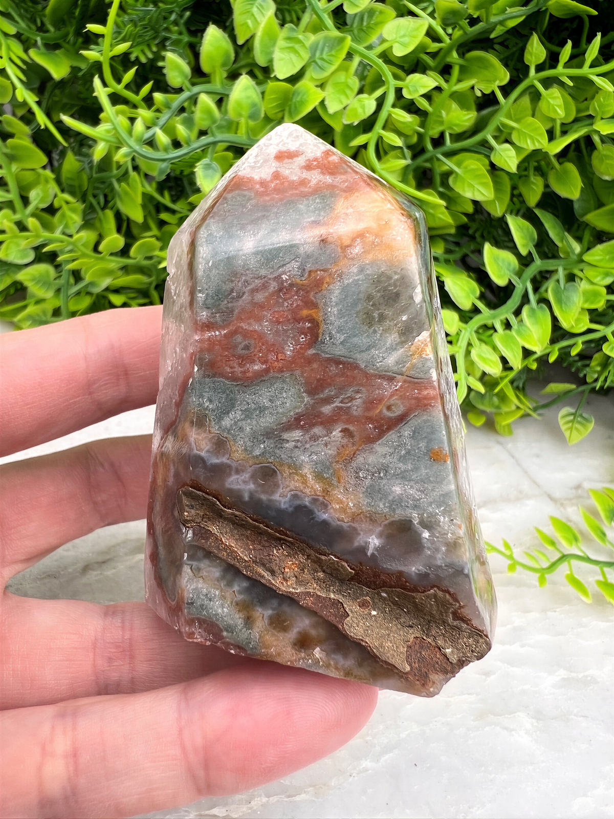 A hand is holding a jagged, multicolored crystal or gemstone against a backdrop of green foliage.