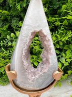 A white geode with a large hole in the center, displayed on a gold stand in front of a background of green foliage.