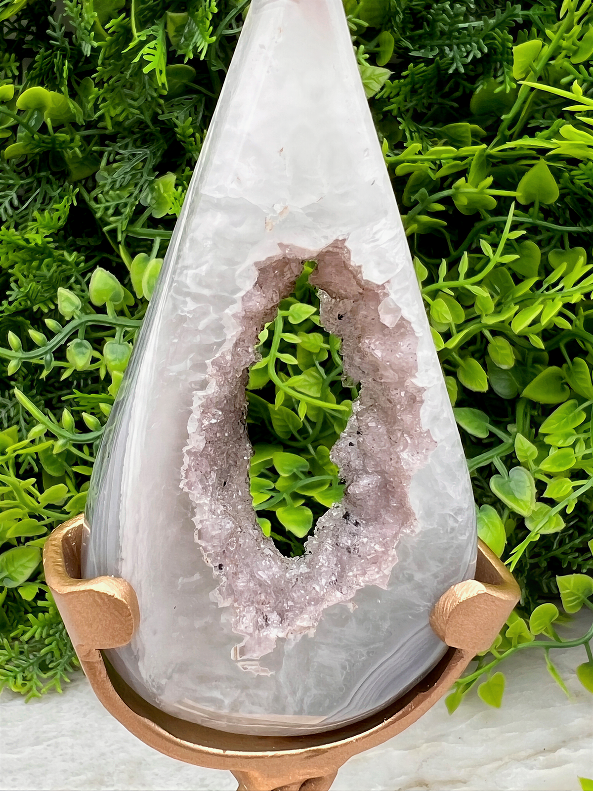 A white geode with a large hole in the center, displayed on a gold stand in front of a background of green foliage.