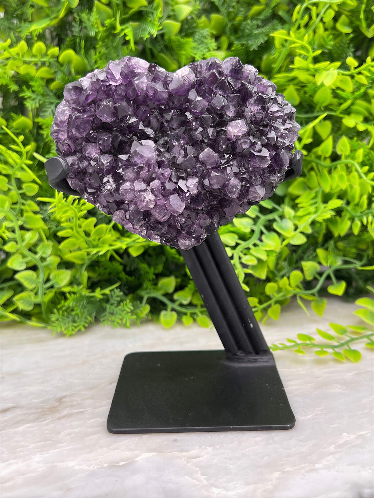 A heart-shaped cluster of purple amethyst crystals is displayed on a black stand, surrounded by lush green foliage.