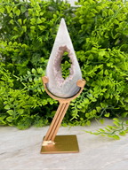 A white geode with a purple interior sits on a gold stand, surrounded by lush green foliage.