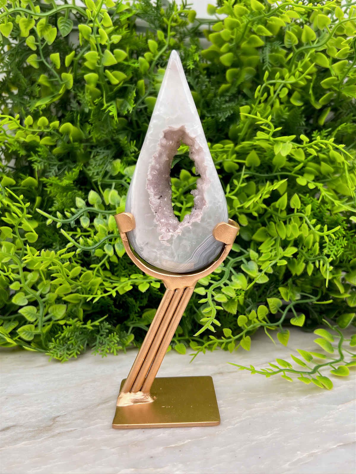 A white geode with a purple interior sits on a gold stand, surrounded by lush green foliage.