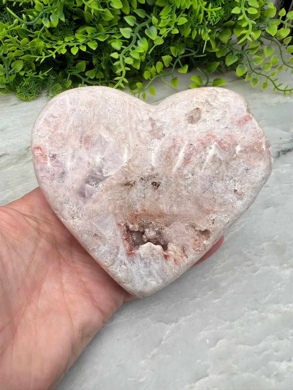 A hand is holding a heart-shaped rock with a pinkish-brown hue, set against a backdrop of green foliage.