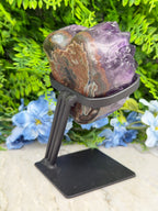 a large purple crystal on a black stand, surrounded by a variety of green plants and blue flowers.