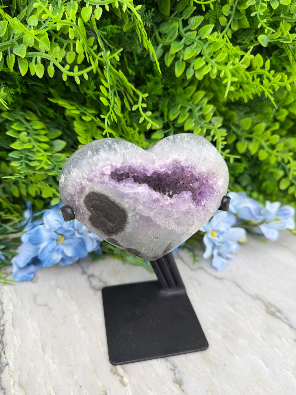 A heart-shaped amethyst geode is displayed on a black stand, surrounded by lush green foliage and blue flowers.