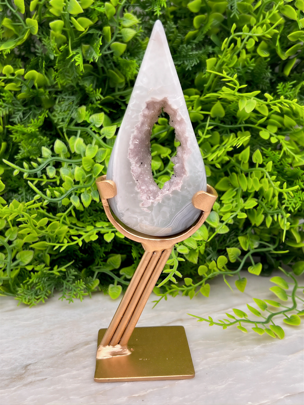 A white geode with a pinkish hue is displayed on a gold stand, surrounded by lush green foliage.