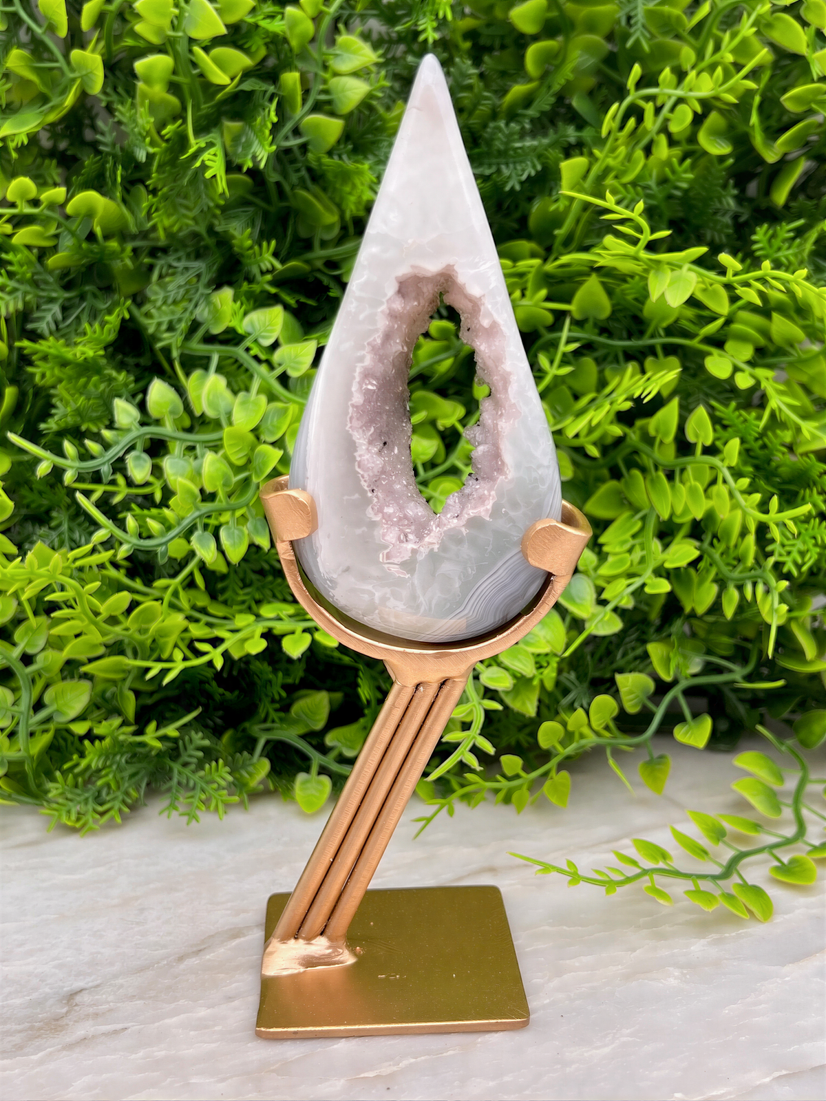 A white geode with a pinkish hue is displayed on a gold stand, surrounded by lush green foliage.