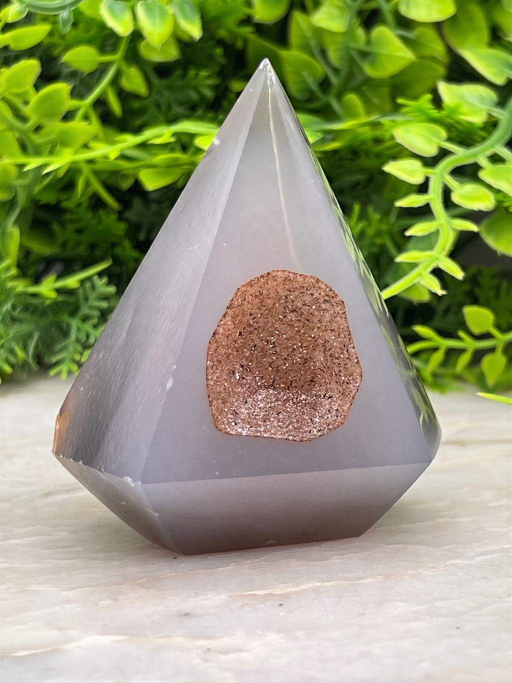 A gray, triangular crystal with a brown, speckled surface sits on a white surface, surrounded by lush green foliage.