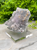 a clear glass display case containing a large, irregularly shaped crystal with a purple hue. The crystal is placed on a marble surface, with a blurred green plant in the background.