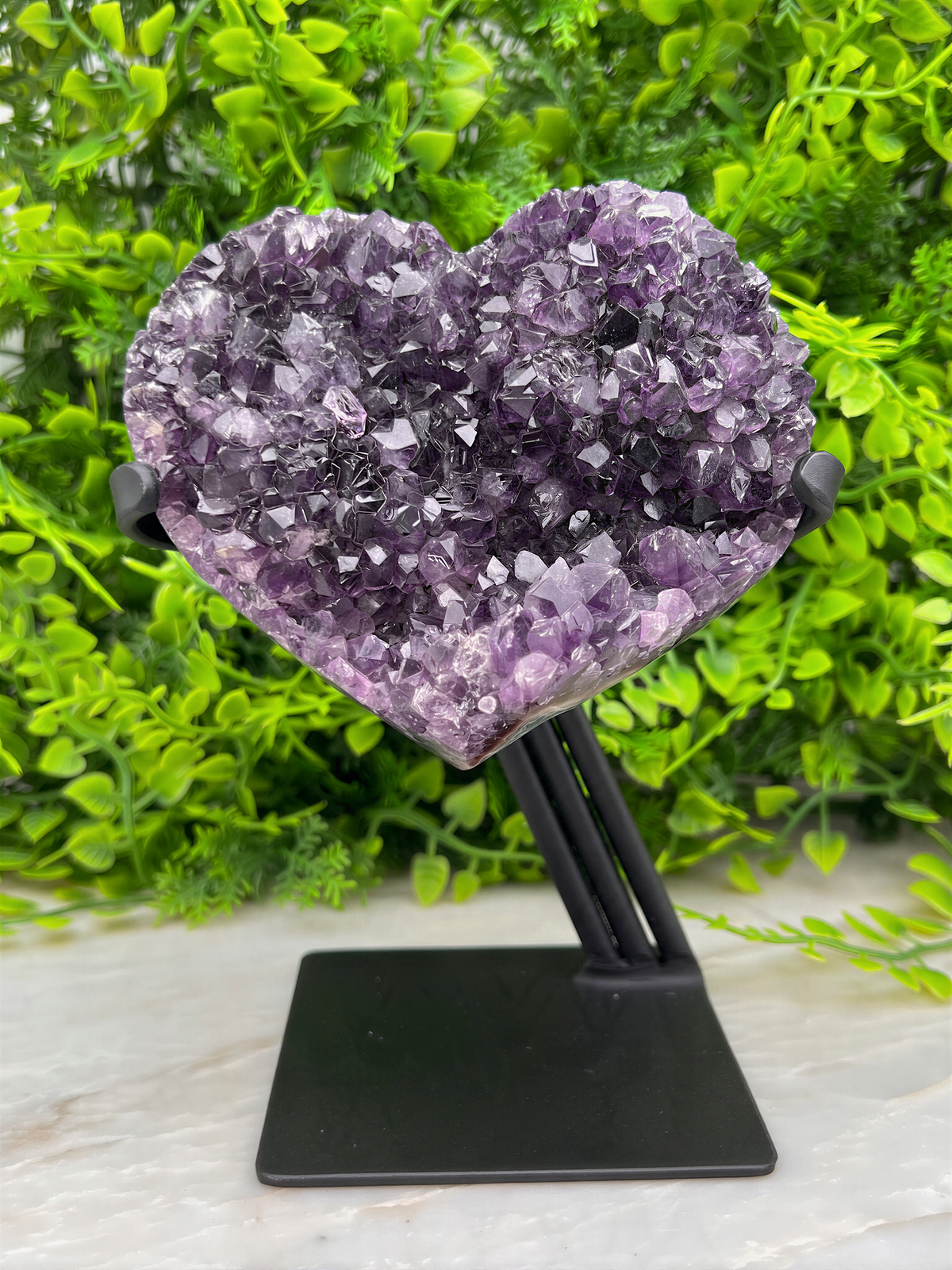 A heart-shaped amethyst crystal rests on a black stand, surrounded by lush green foliage.